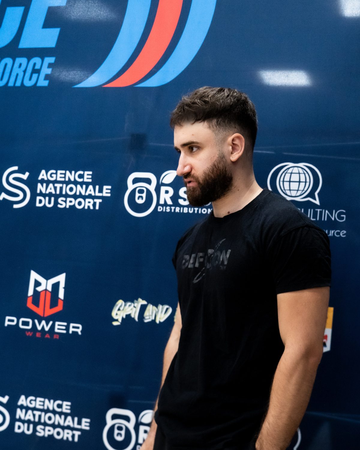 Julien Dinolfo, en t-shirt DEFCON, devant le mur des partenaires d’une compétition FFForce.